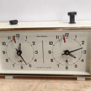 May include: Cream-colored vintage chess clock with two circular clock faces. The clock has black hands, red second hands, and a gold-colored frame. The text "lantaa" and "MADE IN USSR" are visible.