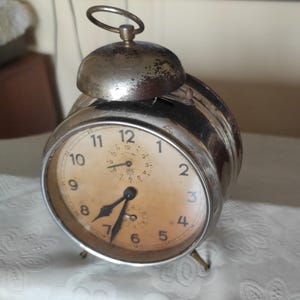 May include: A vintage silver alarm clock with a cream-colored face and black numerals. The clock has a bell on top and stands on small metal legs. The hands point to approximately 6:30.