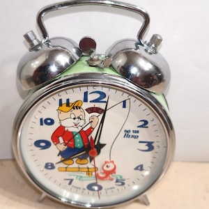 Op de afbeelding: Vintage wekker met een stripfiguur die op de wijzerplaat aan het vissen is. De klok heeft een groene basis, zilveren bellen en een zilveren handvat. De wijzerplaat toont de tijd met zwarte cijfers en wijzers. De tekst "Big Hero" is zichtbaar.