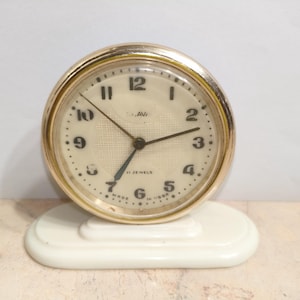 Puede incluir: Un reloj vintage redondo con un bisel dorado y una esfera crema. El reloj tiene números y manecillas negras, y se apoya sobre una base ovalada blanca. La esfera indica "11 JEWELS" y "MADE IN USSR". El diámetro es de aproximadamente 8 cm.