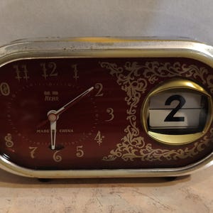 Op de afbeelding: Vintage ovale wekker met een bordeauxrode wijzerplaat en gouden accenten. De klok heeft een analoge weergave met witte wijzers en Arabische cijfers. Een datumweergave toont het nummer 2. De klok is gemarkeerd met "MADE IN CHINA".
