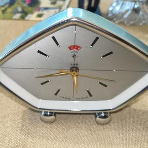 Könnte beinhalten: Ein Vintage-Wecker in Diamantform mit hellblauem Rahmen und silbernem Zifferblatt. Die Uhr hat schwarze Stundenmarkierungen, goldene Zeiger und ein rot-weißes Polaris-Logo. Sie hat zwei verchromte Füße und trägt die Aufschrift "Made in China".