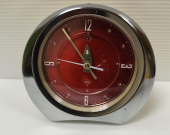 May include: A vintage chrome-cased alarm clock with a vibrant red face. The clock features a diamond-shaped emblem at the bottom and luminous hands. The clock's face displays the numbers 12, 3, 6, and 9.