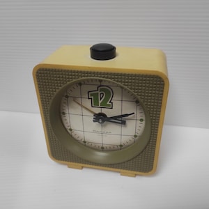 May include: A vintage, square-shaped alarm clock with a cream-colored body and a textured olive green face. The clock face features a grid pattern, a large green number 12, and black hands. A black button sits atop the clock.