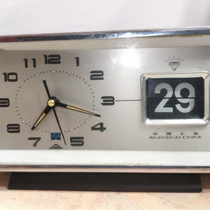 Könnte beinhalten: Analog-Uhr mit silbernem Rahmen, silbernem Zifferblatt, schwarzen Zeigern und goldenen Akzenten. Die Uhr zeigt das Datum 29 in einem Flip-Style-Fenster an. Der Text "SHANGHAI CHINA" ist sichtbar.