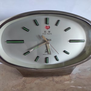 Könnte beinhalten: Ovale silberfarbene Uhr mit hellgrauem Zifferblatt. Die Uhr hat schwarze und grüne rechteckige Stundenmarkierungen und Zeiger. Das Zifferblatt trägt den Text "FIVE RAMS" und ein Logo. Die Uhr steht auf einem silberfarbenen Sockel.