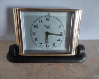 May include: A vintage rectangular clock with a gold-colored frame and a light-colored face. The clock face features black numerals, a small emblem, and hands indicating the time. The clock sits on a black base.