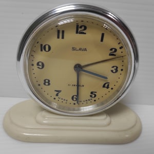 Puede incluir: Un reloj despertador vintage de color crema con un bisel plateado. La esfera del reloj es de color amarillo claro con números y manecillas negros. El reloj indica "SLAVA" y "11 JEWELS". El reloj se encuentra sobre una base crema a juego.