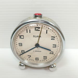 Puede incluir: Un reloj despertador plateado vintage con esfera crema. El reloj tiene números negros, manecillas azules y la palabra "SLAVA" impresa en la esfera. Un botón rojo está en la parte superior. El reloj tiene dos pequeños pies plateados.