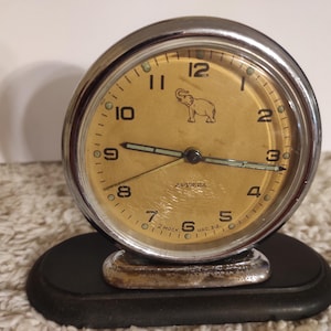 Può includere: Sveglia vintage con quadrante color oro e cornice in metallo argentato. Il quadrante presenta numeri neri, un'immagine di un elefante e la scritta "DRUZHBA". Poggia su una base nera.