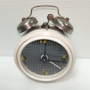Peut inclure: Réveil blanc vintage avec un cadran à motifs noir et blanc et des chiffres jaunes. La montre a deux cloches argentées sur le dessus et une poignée en métal. Les mots "MADE IN USSR" sont visibles sur le cadran.