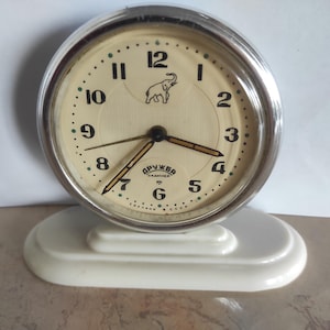 Può includere: Una sveglia vintage color crema con lunetta argentata e base bianca. Il quadrante presenta numeri neri, un emblema di elefante e il testo "ДРУЖБА" e "Сделано в СССР". Le lancette sono nere e dorate.