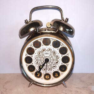 May include: A gold-toned metal twin-bell alarm clock with a white face featuring black circular hour markers numbered one through twelve. The centre of the clock face displays intricate black filigree patterns and twelve small zodiac symbols.