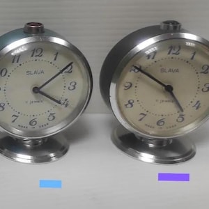 May include: Two round silver-toned metal alarm clocks with circular bases. Each clock features a cream-coloured face with black Arabic numerals, black hands, and the text SLAVA, 11 jewels, and MADE IN USSR printed on the dial. A small button sits on top.