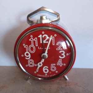 Op de afbeelding: Een vintage rode wekker met een zilveren handvat en poten. De klok heeft een rode wijzerplaat met witte cijfers en wijzers. De tekst "СДЕЛАНО В СССР" staat onderaan de wijzerplaat.