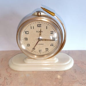 May include: A round Slava alarm clock with a gold-toned metal frame and a cream-colored face featuring black numerals. The clock is mounted on an off-white oval base and includes a gold-toned pointer arm extending from the top of the clock casing.