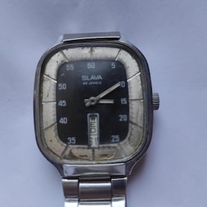Puede incluir: Un reloj de pulsera plateado vintage con esfera negra. El reloj tiene una esfera rectangular con esquinas redondeadas, que muestra la hora y el día. La esfera del reloj tiene la palabra "SLAVA" y "26 JEWELS" impresas. La correa del reloj es de metal plateado.