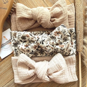 Könnte beinhalten: Drei Baby-Stirnbänder in neutralen Farbtönen. Eines ist einfarbig beige, ein anderes hat ein Blumenmuster mit braunen und cremefarbenen Blumen, und das dritte ist einfarbig hellbeige. Die Stirnbänder sind aus weichem Stoff.