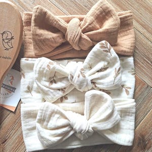 May include: Three fabric headbands with knotted bow details. The top headband is solid tan, the middle one is white with a brown leaf pattern, and the bottom one is solid cream. Each headband features a soft, textured muslin-like fabric construction.