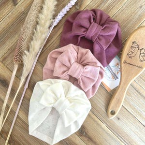 Peut inclure: Trois bandeaux pour bébés, de couleurs mauve, rose et crème. Chaque bandeau est orné d'un grand nœud et d'un motif ajouré. Une brosse en bois avec une illustration de pingouin et du texte est également visible.