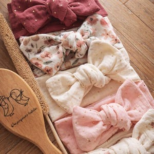 Pode incluir: Uma coleção de faixas de cabelo para bebés em várias cores, incluindo rosa empoeirado, creme e estampados florais. Algumas faixas têm detalhes de pontos dourados. Também está presente uma escova de madeira com uma ilustração de pássaros e texto.