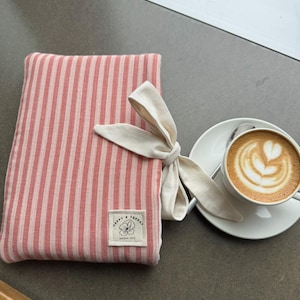 May include: A rectangular fabric cover featuring vertical coral and light pink stripes, accented with a cream-coloured fabric bow tied on the side. A small square label is stitched to the front, displaying the text Poppy & Thread with a flower icon.