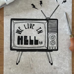 May include: Black and white linocut print of a vintage television with the words "WE LIVE IN HELL" on the screen. The print includes lightning bolt graphics and a skull. A tube of black ink and a carving tool are visible.