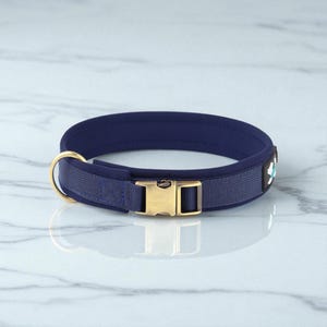 Puede incluir: Un collar para perros azul marino con una hebilla y un anillo de metal dorado. El collar tiene un interior acolchado y un exterior tejido. Un pequeño elemento decorativo con un diseño blanco y turquesa es visible.