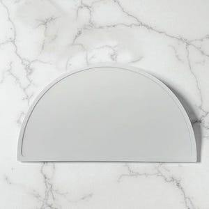 May include: A pale grey, semi-circular silicone mat with a raised edge. The mat has a smooth surface and is shaped like a half-circle. It is likely used in a kitchen or dining setting.
