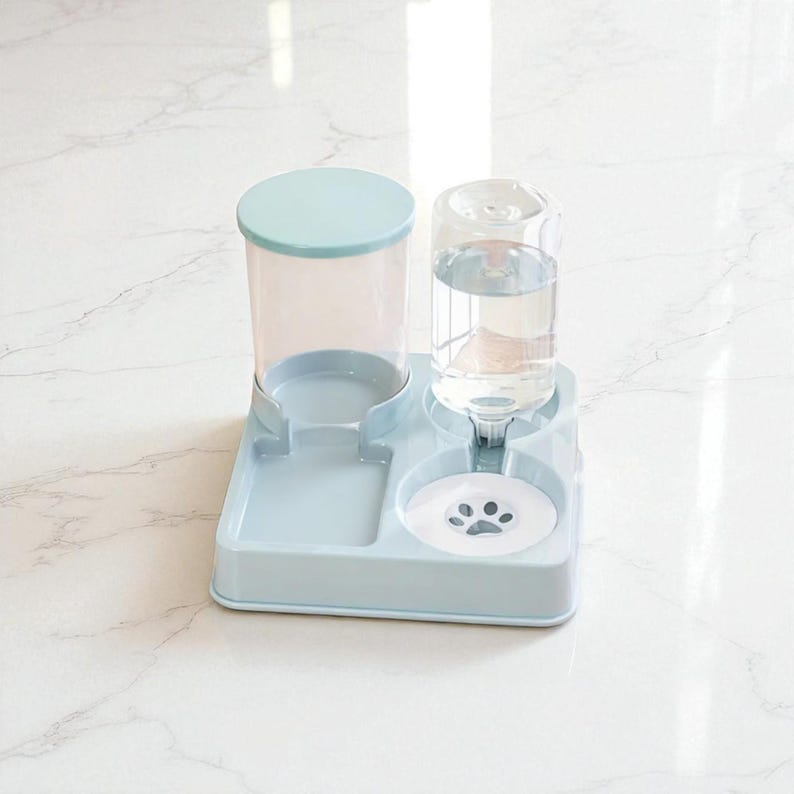 May include: A light blue pet feeder and water dispenser. The water dispenser has a clear, cylindrical bottle filled with water. The feeder has a food bowl and a water bowl with a paw print design. The dispenser has a light blue lid.