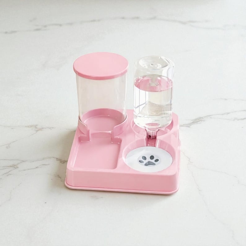 May include: A pink pet water and food dispenser. The dispenser has a clear water bottle, a food container, and a feeding bowl with a paw print design. The dispenser is made of plastic and is designed for small pets.
