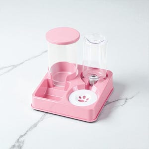 May include: Pink pet food and water dispenser. The dispenser has a square base with a water bottle and a food container. The water bottle is clear plastic and the food container has a pink lid. The base has a paw print design.