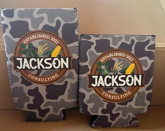 Puede incluir: Dos enfriadores de latas de espuma con estampado de camuflaje gris y negro, con un logotipo circular que incluye el texto "ESTABLISHED 2022 JACKSON CONSULTING" y una ilustración de maíz y hojas verdes. Los enfriadores tienen alturas diferentes.