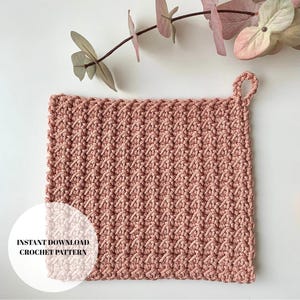 Może przedstawiać: Kwadratowy, pudrowo różowy szydełkowy podkładka pod garnek z teksturowanym wzorem i pętelką do zawieszania. Słowa "INSTANT DOWNLOAD CROCHET PATTERN" znajdują się w białym okręgu w lewym dolnym rogu. Gałązka różowych kwiatów znajduje się w tle.
