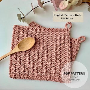 May include: Two square, dusty pink crocheted dishcloths with a textured stitch pattern and small corner loops. A light-colored wooden spoon rests on top. Text on the image reads: English Pattern Only US Terms and PDF PATTERN INSTANT DOWNLOAD.