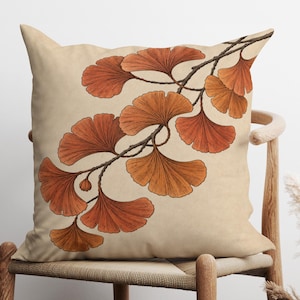 Op de afbeelding: Een vierkant sierkussen met een crèmekleurige achtergrond voorzien van een botanische illustratie van een tak met waaiervormige ginkgobladeren in tinten van gebrand oranje en roestbruin, getekend met fijne donkere lijnen en gedetailleerde nerven.