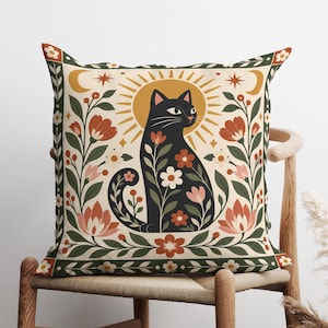 May include: A square throw pillow featuring a black cat illustration centered against a golden sunburst. The cat is surrounded by floral motifs in shades of red, pink, and green. The design includes crescent moons and stars within a decorative border.