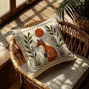 May include: Square decorative pillow with a fox illustration. The pillow features a cream background with a red fox, a red sun, and green foliage. The pillow is placed on a wicker chair.