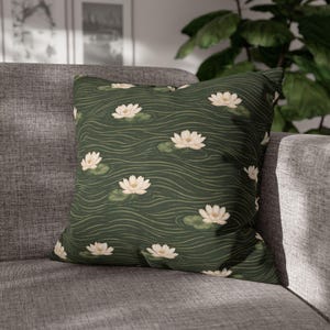 May include: Square decorative pillow with a dark green background featuring a pattern of white water lilies and green lily pads. The pillow is placed on a gray sofa, creating a contrast of colors and textures.