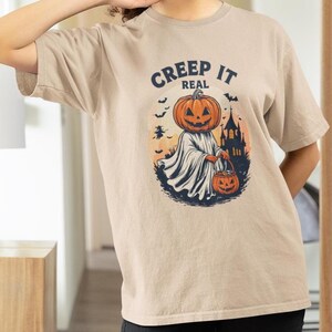 Op de afbeelding: Beige T-shirt met een Halloween-thema. Het ontwerp toont een pompoenhoofdige figuur in een wit laken met een jack-o'-lantern emmer, met de tekst "Creep It Real" erboven. Een spookachtig kasteel en vleermuizen zijn op de achtergrond.