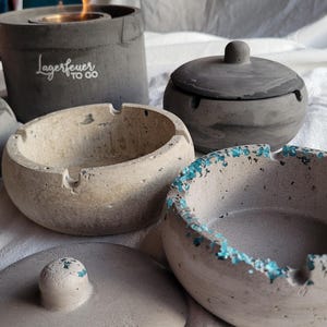 Könnte beinhalten: Eine Sammlung von Aschenbechern und einem kleinen Feuerkorb aus Beton. Die Aschenbecher sind rund mit einer Rille für Zigaretten, einige mit türkisfarbenem Rand. Der Feuerkorb ist zylindrisch mit dem Text "Lagerfeuer to go".