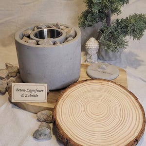 Könnte beinhalten: Ein grauer Beton-Feuerkorb mit Edelstahleinsatz, umgeben von Kieselsteinen. Eine Holzscheibe, ein kleiner Bonsai-Baum und eine Buddha-Statue sind ebenfalls vorhanden. Das Bild enthält ein Schild mit der Aufschrift "Beton-Lagerfeuer & Zubehör".
