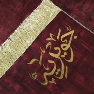 Könnte beinhalten: Ein burgunderfarbener Gebetsteppich mit goldenen Fransen und goldener arabischer Kalligraphie. Die Kalligraphie lautet "Allah ist der Größte".