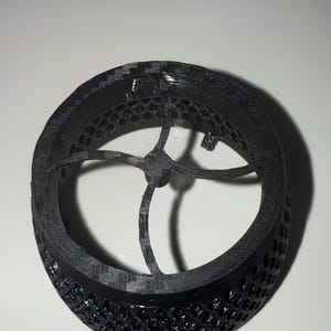 May include: A black 3D printed ring with a honeycomb pattern and a four-spoke design in the centre.