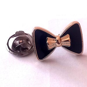 May include: A navy blue bow tie lapel pin with a gold-coloured centre and trim. The pin has a classic bow tie shape and a silver-coloured pin back. This accessory is suitable for formal wear.