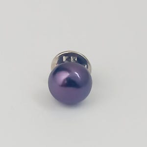 May include: A single, round, dark purple pearl lapel pin with a silver-colored backing. The pearl has a glossy finish, reflecting light. The pin is set against a plain white background, highlighting the jewelry.