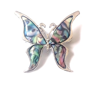 Può includere: Spilla a farfalla argentata con ali iridescenti. Le ali presentano un mix di blu, verdi e viola, con un contorno argentato. La spilla ha un piccolo accento di cristallo al centro.