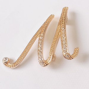 Może przedstawiać: Złota broszka z metalu w kształcie litery "M", ozdobiona przezroczystymi kryształkami. Broszka ma kursywny design z pojedynczym kryształkiem u podstawy litery. Broszka jest umieszczona na białym tle.