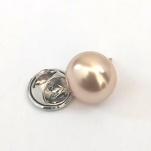 May include: A close-up of a pearl lapel pin. The pin features a lustrous, light-pink pearl attached to a silver-colored metal backing. The pin is designed to be attached to clothing or accessories.