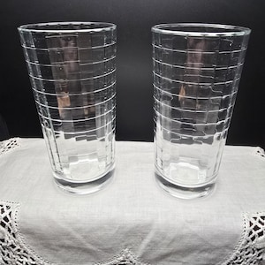 May include: Two clear, tall drinking glasses with a grid pattern. The glasses are made of glass and sit on a white, lace-edged cloth. The background is black.